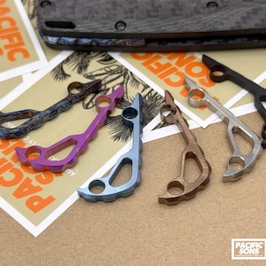Puede incluir: Una colección de pernos de pulgar para cuchillos personalizados y coloridos en varios colores, incluyendo morado, azul y bronce. Los pernos se muestran sobre una superficie beige con la marca Pacific Sons.