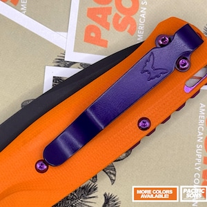 Può includere: Coltello tascabile arancione con lama nera e dettagli viola. Il coltello presenta una clip da tasca viola con logo a farfalla e viti viola. La lama è contrassegnata con "S30V". Il coltello è posto su uno sfondo di biglietti stampati.
