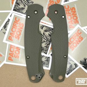May include: Two green textured knife scales with rounded edges and multiple holes. The scales are on a brown surface with several white squares with the text "Pacific Sons American Supply Company" printed on them.