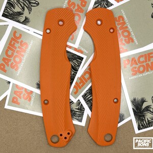 May include: Two orange knife scales with a textured surface. The scales have a rounded shape and are designed to fit a knife handle.