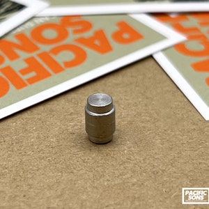 Puede incluir: Un botón pulsador de metal plateado con una parte superior lisa y redondeada. El botón mide aproximadamente 2,5 cm de alto y tiene una superficie ligeramente texturizada.