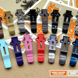 May include: A collection of colorful pocket clips in various finishes, including brown, gold, black, and patterned designs. The clips are arranged on a surface with the text "PACIFIC SONS" and "AMERICAN SUPPLY COMPANY". The image also includes a banner that says "MORE COLORS AVAILABLE!"