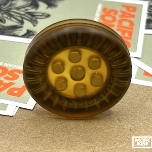 May include: A circular, translucent amber-colored button featuring a raised central hub with six small, evenly spaced circular protrusions. The outer rim of the button has a series of vertical, grooved ridges that extend around the entire circumference.
