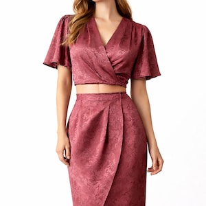 May include: A two-piece formal outfit in a dusty rose color. The top is a wrap-style with short sleeves. The skirt is a tulip style, with a high waist and a knee-length hem. Both pieces have a subtle floral pattern.
