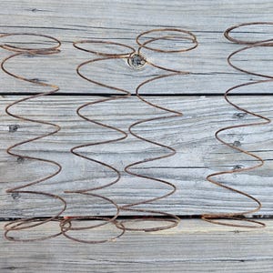 May include: Three rusty metal bed springs arranged on a weathered wooden surface. The springs have a zigzag pattern with circular ends, showcasing a vintage or industrial aesthetic. The wood has a gray tone.