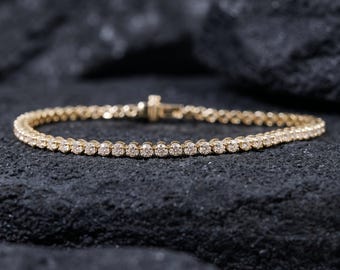 Può includere: Un bracciale tennis in oro caratterizzato da una fila continua di pietre rotonde, trasparenti e sfaccettate, incastonate in una montatura metallica. Il bracciale include una chiusura a cassetta sicura al centro, con un design uniforme di gemme.
