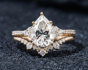 Könnte beinhalten: Ein Ring-Set aus Roségold mit einem zentralen, birnenförmigen, klaren Edelstein, der von einem Kranz aus kleineren, runden, klaren Steinen umgeben ist. Die Ringschiene ist mit winzigen Steinen besetzt, ergänzt durch einen passenden, geschwungenen Ring.
