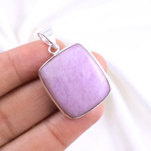 May include: A rectangular pendant with a light purple gemstone set in a silver-colored metal frame. The gemstone has a smooth, polished surface and a subtle, marbled appearance. The pendant has a small loop at the top for a chain.