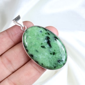 Puede incluir: Un colgante de forma ovalada que presenta una piedra verde pulida con inclusiones negras oscuras, engastada en un marco de metal de tono plateado liso con una anilla triangular en la parte superior para sujetarlo a una cadena, con superficie lisa.
