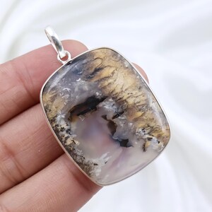 Puede incluir: Un colgante rectangular con una piedra preciosa de ágata dendrítica pulida engastada en un marco de plata liso. La piedra muestra tonos blancos y grises translúcidos con intrincadas inclusiones negras y marrón dorado en forma de helecho.