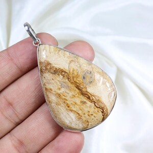 May include: A teardrop-shaped pendant with a silver-colored bail. The pendant features a polished, light beige stone with brown veining and a silver-colored bezel. The stone has a unique, natural pattern.