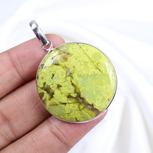 Peut inclure: Un pendentif rond avec une pierre vert citron vif sertie dans un cadre en métal argenté. La pierre présente un motif naturel unique avec des inclusions vertes plus claires et brunes. Le pendentif a une petite boucle en haut pour une chaîne.