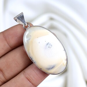 Könnte beinhalten: Ein ovaler Anhänger mit einem glatten, polierten cremefarbenen Stein, der dezente beige Wirbel und dunkle dendritische Einschlüsse aufweist. Der Stein ist in eine schlichte silberfarbene Metallfassung mit einer dreieckigen Öse eingefasst.