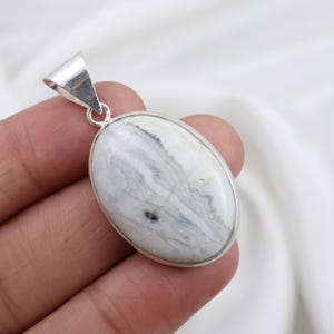 May include: Oval-shaped pendant with a polished, marbled stone in shades of white, gray, and pale blue, set in a silver-tone metal frame. The pendant has a simple bail for attaching to a chain. The stone's pattern resembles flowing water.