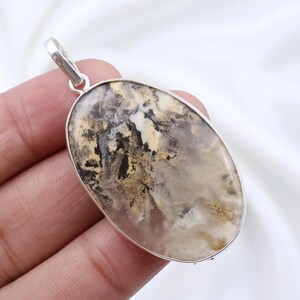 Puede incluir: Un colgante de forma ovalada con una piedra pulida y translúcida que presenta intrincadas inclusiones naturales en tonos crema, beige, marrón oscuro y negro. La piedra está engastada en un bisel de metal plateado liso con una anilla a juego.