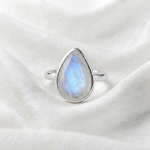 Puede incluir: Un anillo de plata con una piedra de luna en forma de pera, de aspecto translúcido y con un suave brillo azul iridiscente. La gema está engastada en un bisel de plata lisa y pulida, unido a una banda de plata sencilla y delgada.
