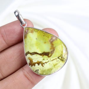 Peut inclure: Un pendentif en forme de goutte d'eau avec une bélière argentée. Le pendentif présente une pierre vert clair avec des veines marron et beige. La pierre est sertie dans une lunette argentée. Le pendentif est tenu dans une main.