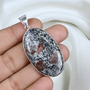Peut inclure: Pendentif ovale avec une bélière argentée. Le pendentif présente une pierre mouchetée grise et blanche avec des inclusions brun rougeâtre. La pierre est sertie dans une lunette argentée.