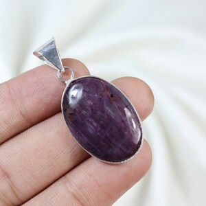 Könnte beinhalten: Ovaler Anhänger mit einem tiefvioletten Edelstein. Der Stein ist in einen silberfarbenen Metallrahmen gefasst und hat eine polierte Oberfläche. Der Anhänger hat eine Schlaufe für eine Kette und einen dreieckigen Bügel.