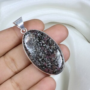 以下が含まれることがあります： 銀色の金属フレームにセットされた、斑点のある濃いグレーとピンクの石を使用した楕円形のペンダント。ペンダントの上部にはチェーン用のループが付いています。