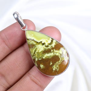 Peut inclure: Un pendentif en forme de goutte d'eau avec une bélière en argent. Le pendentif présente une pierre précieuse polie avec un mélange de teintes jaunes, marron et vertes. La pierre est sertie dans une lunette en argent.