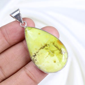 Peut inclure: Pendentif en forme de poire avec une pierre précieuse vert clair, veinée de brun et de jaune. La pierre est sertie dans un cadre en métal argenté et possède un bélière argentée pour une chaîne. Le pendentif est tenu dans une main.