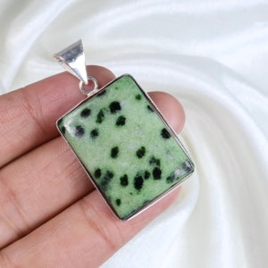 Può includere: Un ciondolo rettangolare caratterizzato da una pietra verde chiaro con inclusioni maculate verde scuro e nere, incastonato in una cornice di metallo color argento lucido con un gancio triangolare sulla parte superiore.