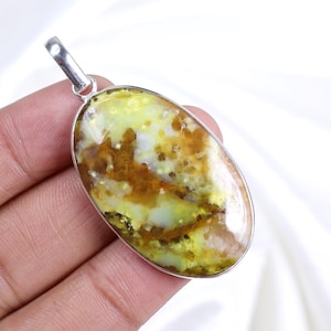 Peut inclure: Pendentif ovale avec une bélière argentée. Le pendentif présente une pierre polie avec un mélange de couleurs jaune, marron et blanc. La pierre est sertie dans un cadre en métal argenté. Le pendentif est tenu dans une main.