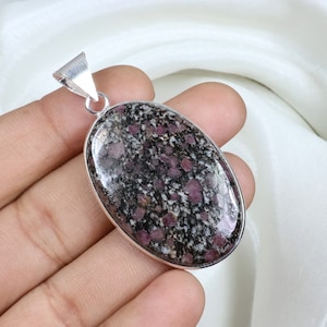 Könnte beinhalten: Ovaler Anhänger mit einem gesprenkelten, dunkelgrauen und rosa Edelstein, der in einen silberfarbenen Metallrahmen gefasst ist. Der Anhänger hat eine Öse zum Anbringen einer Kette. Der Edelstein hat ein einzigartiges, natürliches Muster.