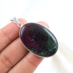 May include: Oval-shaped pendant with a silver-colored bail. The pendant features a polished gemstone with a mix of deep red, green, and black colors. The gemstone is set in a silver bezel.