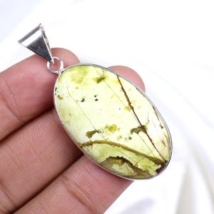 Peut inclure: Pendentif ovale avec une pierre vert clair, présentant des veines marron et vert foncé. La pierre est sertie dans un cadre en métal argenté avec une bélière assortie. Le pendentif mesure environ 5 cm de long.