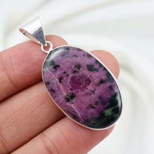 Op de afbeelding: Een ovaalvormige hanger met een gepolijste edelsteen met een gevlekt patroon van diep magenta, zwarte en donkergroene insluitingen. De steen is gezet in een gladde, zilverkleurige metalen rand met een bijpassende driehoekige hanger aan de bovenkant.
