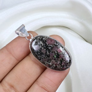 May include: Oval pendant with a dark, speckled stone, displaying red and green inclusions, set in a silver-coloured metal frame. The pendant has a loop at the top for a chain. The stone's surface is polished and reflective.