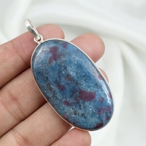 May include: Oval-shaped pendant with a blue and red stone set in a silver-colored metal frame. The stone has a mottled appearance with dark blue and red hues. The pendant has a loop at the top for a chain.