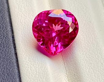 Puede incluir: Una gema facetada en forma de corazón, de un tono rosa vibrante. La gema se presenta sobre un fondo blanco y gris, resaltando su talla brillante y sus superficies reflectantes. El color de la gema es un rosa rico y saturado.