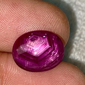 Puede incluir: Primer plano de una gema pulida, de forma ovalada y de color magenta intenso. La gema tiene un complejo patrón interno de líneas rosas claras y oscuras, creando un efecto de estallido estelar. La piedra se sostiene entre los dedos.