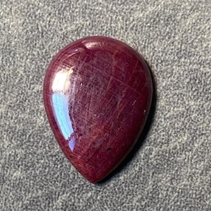 Könnte beinhalten: Ein tiefroter Edelstein-Cabochon, geschliffen in Tropfenform. Der Stein hat eine glänzende Oberfläche und ist vor einem grauen Hintergrund platziert.