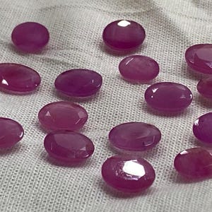 May include: A selection of oval and round-cut, faceted ruby gemstones. The rubies are a deep pinkish-red colour and are displayed on a white, textured fabric. The gemstones vary in size and shape, with a polished, reflective surface.
