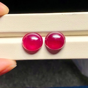 Könnte beinhalten: Zwei runde, im Cabochon-Schliff geschliffene Edelsteine mit einer tiefen, leuchtend roten Farbe und einer glatten, polierten Oberfläche. Diese Steine zeichnen sich durch eine transluzente Qualität aus, die das Licht einfängt und Reflexe erzeugt.