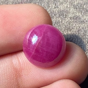 Puede incluir: Una piedra preciosa de rubí pulida y redonda. La gema es de color rojo rosado intenso con una superficie lisa y reflectante. La piedra preciosa se sostiene entre dos dedos.
