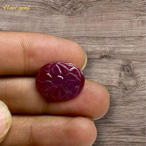 Op de afbeelding: Een ronde, gepolijste robijn edelsteen met een gesneden bloemenpatroon. De dieprode steen wordt tussen vingers gehouden, wat de ingewikkelde details laat zien. De Etsy shop naam "Hoot gems" is zichtbaar.