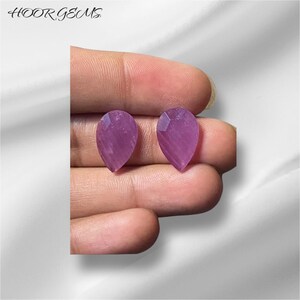 Pode incluir: Duas pedras preciosas em forma de pêra, cada uma com uma tonalidade roxa e um corte facetado. As pedras preciosas são mantidas em uma mão.