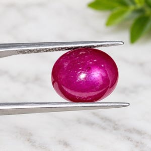 Op de afbeelding: Een levendige, ovaalvormige, diep roze-rode edelsteen met een glad, gepolijst cabochon oppervlak. De steen wordt stevig vastgehouden tussen de twee metalen punten van een zilverkleurige pincet, wat de doorschijnende en reflecterende kwaliteit toont.