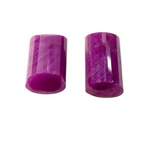 Op de afbeelding: Twee gefacetteerde, rechthoekige, diep magenta edelstenen. De edelstenen hebben een gepolijst, reflecterend oppervlak en staan tegen een witte achtergrond. De tekst "Hoot gems" staat in de linkerbovenhoek.