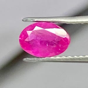 Peut inclure: Une pierre précieuse vibrante de taille ovale avec une couleur rose profonde et saturée. La pierre présente de multiples facettes triangulaires et en forme de cerf-volant sur sa surface, créant un aspect brillant et réfléchissant. Elle est tenue par des pinces en métal argenté.