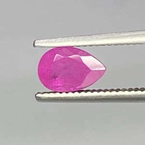 Può includere: Una pietra preziosa sfaccettata a forma di pera con una vibrante tonalità rosa. La pietra è tenuta saldamente tra le due punte metalliche seghettate di una pinzetta, mettendo in risalto la sua qualità traslucida e il taglio geometrico.