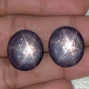 May include: Two round, polished, star sapphire gemstones. The stones are a deep purple color with a shimmering star effect visible across their surfaces. The gemstones are held in a hand.