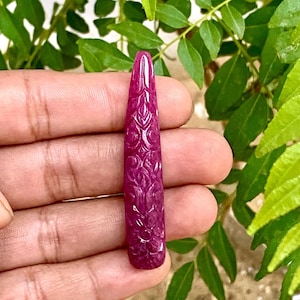 Könnte beinhalten: Ein geschnitzter, länglicher, tiefmagentafarbener Edelstein mit aufwendigen floralen Mustern. Der Edelstein wird vor einem Hintergrund aus grünen Blättern gehalten, was seine satte Farbe und detaillierte Handwerkskunst zeigt. Der Edelstein ist etwa 7.6 cm lang.