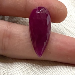 Puede incluir: Una gema de rubí facetada en forma de lágrima. La piedra de color rojo púrpura oscuro se sostiene entre los dedos, mostrando sus superficies pulidas y reflectantes. La forma alargada de la gema se estrecha hasta un punto, con múltiples facetas que capturan la luz.