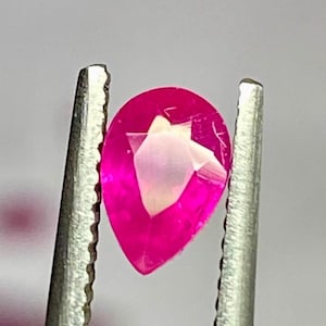 Op de afbeelding: Een levendige, peervormige losse edelsteen met een diepe, verzadigde fuchsia roze kleur en meerdere gefacetteerde oppervlakken die licht reflecteren, stevig vastgehouden tussen de gekartelde metalen punten van een zilverkleurige pincet.